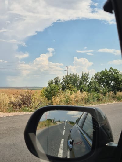 The image captures a view from inside a car, looking out the passenger side window and also at the side mirror. The scene depicts a rural road with dry vegetation and trees lining the right side of the asphalt road. In the distance, a flat, open landscape stretches out under a bright blue sky filled with fluffy white clouds. A utility pole with wires is visible on the left side of the frame, suggesting the presence of infrastructure in this rural area.
The side mirror reflects the road ahead, showing the car's path, which appears to be a straight stretch of highway. There are traffic signs visible in the reflection, including a circular sign with "50" inside a red circle, indicating a speed limit. The sunlight suggests it is daytime, likely late morning or afternoon, with clear and pleasant weather. The dry, straw-like vegetation points to a dry season, possibly summer or early autumn. The overall mood is one of travel and observation, capturing a moment on a road trip through a sunny, rural Bulgarian landscape.