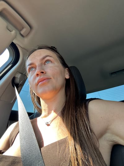 The image shows a woman sitting in the driver's seat of a car, looking upwards and to the left. She has long brown hair and blue eyes. She is wearing a dark brown top and a silver necklace with a single gemstone pendant. A seatbelt is visible across her chest. The interior of the car is visible, including the ceiling and a sun visor. The lighting suggests it is daytime, with sunlight entering from the left side of the car. The background appears to be an outdoor environment with a clear blue sky. The location is not discernible from the image itself.