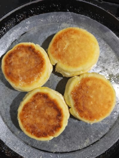 In a grey, circular frying pan with a black rim, four golden-brown, round, pancake-like pastries are cooking. They are arranged in a diamond formation, with one at the top, one at the bottom, and two on either side. The pastries have a slightly uneven, textured surface with some areas appearing lighter yellow and others a deeper golden brown, suggesting they are being fried. Small white flecks, possibly salt or sugar, are visible on the surface of some of the pastries. The frying pan appears to be on a stovetop, as indicated by the black, textured edge of the pan. The background is dark and out of focus. The scene is located in Dolzhikovo, Russia.