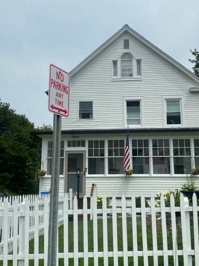 The image depicts a white, two-story residential structure located at 4 Catoonah Street, Ridgefield, Connecticut, United States, under an overcast sky. In the foreground, a white wooden picket fence separates a green lawn from the viewer's perspective. To the left of center, a grey metal pole supports a red and white rectangular 'NO PARKING ANY TIME' sign with double-headed horizontal arrows. The house features horizontal white siding and a dark gabled roof. The first story presents an enclosed porch with multiple large windows, above which the word 'Four' is displayed. A screened entry door and a black outdoor lamp are visible on the porch. An American flag hangs vertically from the porch ceiling, right of center. Two hanging basket planters are suspended from the porch overhang. The second story has three rectangular windows, while the gable end features a central arched window flanked by two narrow rectangular windows, with a small rectangular vent above. An indistinct dark shape, consistent with a dog's head, is visible looking out from the far-right window of the enclosed porch. Dark green foliage is present to the left and behind the house, and lighter green plants with white flowers are in a garden bed to the right of the porch.