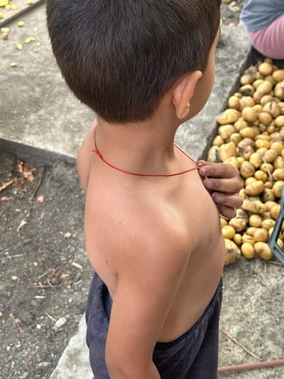 The image, taken in Mlekarevo, Bulgaria, shows a close-up, top-down view of a young person's upper body and head. The person, whose face is not visible, has dark hair and is wearing dark pants and a red string tied around their neck. A pile of potatoes is visible in the background, suggesting an outdoor agricultural setting. The lighting indicates it is daytime. The person's hand, with dirt on the fingers, is resting near their neck. The scene appears to be outdoors, possibly in a rural area. There is no visible text in the image.
