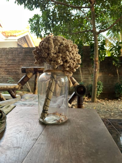 A dried flower arrangement in a clear glass bottle sits on a wooden table outdoors. The bottle is tied with twine and contains a few dried leaves at the bottom. Behind the table, a brick wall is visible, with a rustic wooden bench or chair and greenery. In the background, a tree with green foliage and part of a building with an orange tiled roof are present. The scene suggests a casual outdoor setting, possibly a garden or patio area in Karangkebagusan, Indonesia.