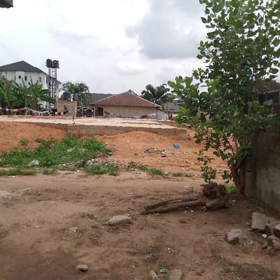 The image shows a dirt lot located in Adanisievbe, Nigeria at the coordinates 5.953 latitude, 5.705 longitude. The lot is in front of a recently constructed building. It is partially covered in dirt and grass, and some of the building's foundation can be seen. A large tree is to the right of the image with its roots exposed. Some trash and debris can be seen in the lot. The building in the background appears to be recently completed and is a single story with a flat roof. There are no people visible in the image.