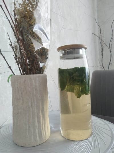 A still life image is presented, likely taken indoors, given the artificial lighting and the presence of a grey upholstered chair in the background. The scene is dominated by two objects on a textured white surface. On the left is a cream-colored ceramic vase with an irregular, textured surface, containing a bouquet of dried flowers and branches, some of which are wrapped in clear plastic. To the right of the vase is a clear glass carafe filled with a pale yellow liquid and what appears to be fresh mint leaves. The carafe has a wooden lid. The background wall has a subtle textured pattern with thin, dark lines. The presence of a chair suggests a domestic or possibly an office setting. The city of Novostroika, Russia is not depicted in this image, as there are no elements within the frame that would indicate its location.