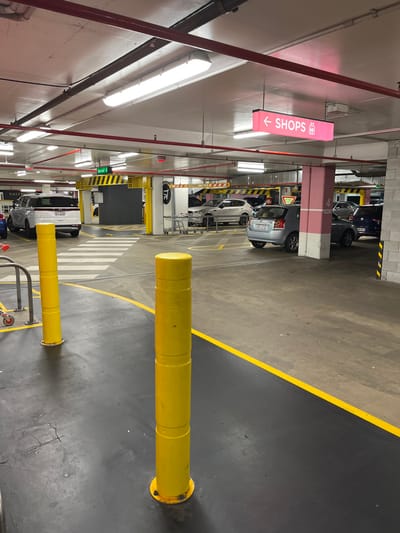 This media file captures an indoor parking garage, likely a multi-story or underground facility, in what appears to be a functional, utilitarian setting. The image is oriented vertically but depicts a scene that would typically be viewed horizontally, with the right side of the image showing the ceiling and the left side showing the floor.

The scene features multiple vehicles, mostly cars, parked and moving within the garage. The floor is a mix of dark grey and lighter concrete sections, clearly delineated by bright yellow lines, indicating parking bays and roadways. A prominent feature on the ground level is a large, yellow, cylindrical bollard in the foreground, with another similar, smaller bollard visible further back. A pedestrian crossing with white stripes is visible in the mid-ground.

Overhead, numerous fluorescent tube lights illuminate the area, and red pipes run along the ceiling. A notable object is a pink, rectangular sign suspended from the ceiling, which reads "SHOPS" with a left-pointing arrow and a pictogram of a person. This indicates a pedestrian route to retail areas. In the background, yellow and black striped barriers, possibly for vehicle access control, are visible, one displaying the letters "TTC". Large pillars, some painted in a reddish-brown hue and others grey, support the structure.

A few people are faintly visible near parked cars or near the access barriers, some wearing face masks, suggesting a contemporary context. The overall impression is one of a busy, operational parking facility, common in commercial or retail centers like those found in Indooroopilly, Australia. There is no indication of time of day or weather as the environment is entirely indoors.