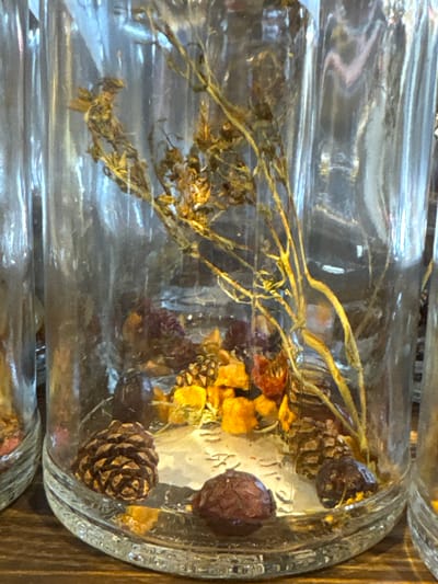 A close-up shot of two glass jars filled with dried botanical elements. The visible contents include dried sprigs, yellow and red flower pieces, and pinecones. The jars appear to be cylindrical, with thick glass and ridges around the top. The background is blurred, but a wooden surface is visible. The image is taken in a natural light setting. The photograph does not depict the city of Kurbaki, Russia.