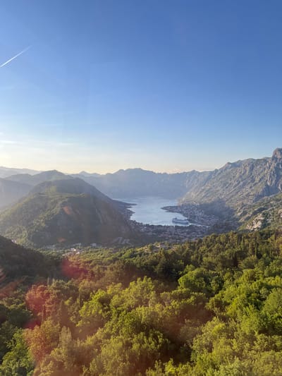 This elevated, wide-angle shot captures the panoramic view over the Bay of Kotor and the city of Kotor, Montenegro, from the mountainous slopes near Čavori. The scene is illuminated by the low sun, suggesting either early morning or late afternoon, with warm light highlighting parts of the landscape and creating lens flare in the lower half of the image.
The upper third of the image is dominated by a clear, gradient blue sky, transitioning from a deeper blue at the top to a paler hue near the horizon. A single, faint white contrail streaks diagonally across the upper left quadrant.
The middle and background feature the expansive Bay of Kotor, a serene body of blue-grey water, winding between imposing, rugged mountains. On the eastern shore of the bay, the densely built city of Kotor is visible, characterized by numerous structures and a large, white cruise ship prominently docked or anchored near its waterfront. Beyond the city, a series of steep, rocky mountain ranges rise dramatically, extending into the atmospheric haze of the distance.
The foreground, comprising the lower third of the image, is covered with a thick canopy of vibrant green trees and lush vegetation, indicative of forested slopes. Sunlight illuminates the treetops, creating bright highlights and contrasting with shadowed areas. A faint, winding road can be discerned among the trees in the mid-foreground, leading towards the distant city. No individuals or specific human activities are discernibly captured within the frame.