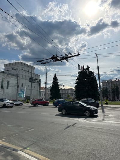 A street scene in a city, likely Putyatino (Semivragovskaya S/A), Russia, on a partly cloudy day. Overhead wires for a trolleybus system crisscross the frame. On the left, a white, fortress-like building with crenellated battlements and arched windows stands prominently. In front of it, several cars are visible on the road: a white SUV, a red SUV, a dark sedan, and a dark blue sedan. A tall evergreen tree is positioned behind the cars, near a lamppost. Further in the background, various multi-story buildings with classical architectural elements line the street. The sky is bright with the sun peeking through scattered clouds, casting shadows on the asphalt road and vehicles. The road has lane markings, including a crosswalk.