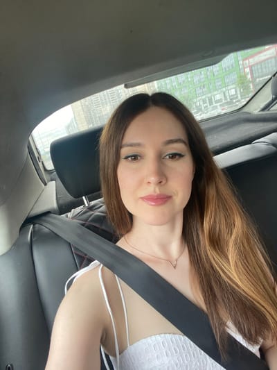A young woman with long, brown and blonde hair is sitting in the passenger seat of a car. She is wearing a white, sleeveless dress with a textured pattern and a simple rose gold necklace. A black seatbelt is fastened across her chest. Her makeup includes winged eyeliner and glossy pink lipstick. The woman is looking at the camera with a slight smile. Through the car window, a blurred view of buildings and trees is visible, suggesting an urban or suburban setting. The lighting indicates it is daytime. The overall mood is casual and relaxed. The location is identified as Колос Снт, Russia.