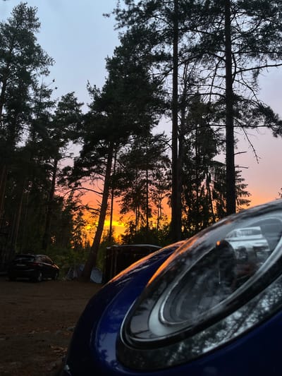 This image captures a campsite scene at dusk in Luzhki, Russia. In the foreground, the curved, blue surface of a car's headlight reflects the warm hues of the setting sun. The sky behind is a gradient of soft grey and vibrant orange, with the sun's brilliant glow visible through a dense forest of tall pine trees. The trees, silhouetted against the bright sky, create a dark canopy. In the mid-ground, a dark-colored SUV is parked on a dirt path. Several tents are set up near the trees, suggesting a camping activity. The ground is covered with dirt and scattered stones. A makeshift structure, possibly a shelter, can be seen towards the right side of the image, adorned with colorful bunting. The overall atmosphere is tranquil, with the natural beauty of the sunset setting a peaceful mood over the campsite.