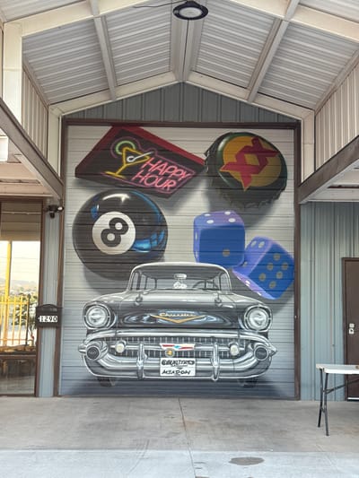 The image shows a garage door in Sunland Park, United States of America, that has been painted with a mural. The mural features a vintage black Chevrolet car in the center, with various objects arranged around it. To the top left of the car, a neon sign displays a cocktail glass and the words "HAPPY HOUR". To the top right, a large bottle cap with a red and yellow starburst design is depicted. Directly above the car, a large black eight-ball from a billiard game is painted. To the right of the eight-ball, two oversized blue dice are shown, one with four dots and the other with five dots. The garage door itself is made of corrugated metal and is painted to appear as a mural. The surrounding environment includes the roof structure of the garage, with exposed beams and corrugated metal roofing. To the left of the garage door, a glass door and a mailbox with the number "1290" are visible. A metal table with a white surface stands to the right of the garage door. The ground in front of the garage is concrete.