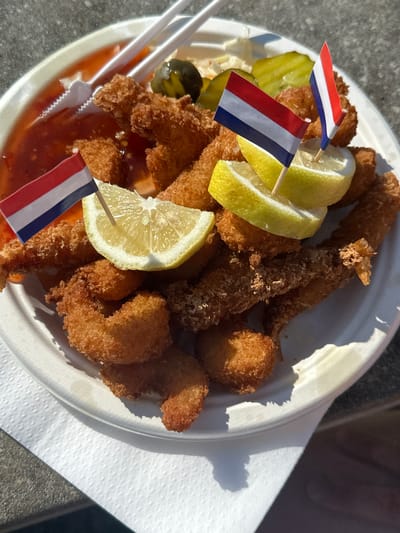A plate of fried shrimp is presented on a white paper plate, garnished with lemon wedges and small Dutch flags. A red dipping sauce is visible on the side of the plate, along with some pickles. The plate is placed on a white paper napkin, which rests on a textured grey surface, possibly a table or counter. The background is slightly blurred, suggesting an outdoor setting. The lighting indicates it is daytime, with sunlight casting shadows. The scene is static, with no people actively interacting with the food. The overall impression is of a casual meal, possibly from a food stall or casual restaurant in Zandvoort, Netherlands, given the presence of the Dutch flags.