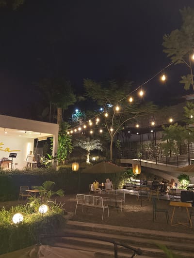 This image depicts an outdoor dining area at night in Caringin, Indonesia. A white building with a stage is visible on the left, where a person is standing. String lights illuminate the scene, casting a warm glow over tables and chairs set up in a gravel-covered courtyard. Several patrons are seated at tables, enjoying their evening. Lush greenery surrounds the seating area, and decorative lamps hang from trees, adding to the ambiance. The overall atmosphere suggests a relaxed and inviting dining experience.