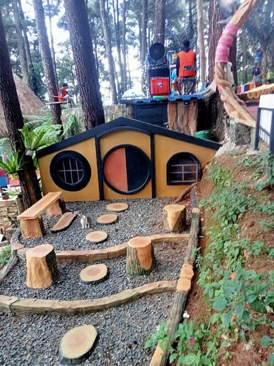 This image depicts a detailed outdoor scene in a themed recreational area within a dense pine forest, specifically located in Kuwukan, Indonesia.

In the foreground and mid-ground, a multi-tiered pathway is visible, surfaced with grey gravel. This path is bordered by textured, dark-brown, imitation-log retaining walls and steps. Circular, light-colored stepping stones, designed to resemble tree cross-sections, are embedded along the path. Several realistic-looking tree stump replicas serve as seating or decorative elements, alongside a single light-colored, simulated wooden bench on the far left. A small, bicolored domestic rabbit, predominantly white with grey/brown markings, stands on the gravel path in front of a structure.

At the center-left, a small, low-profile building with a yellow-ochre facade and dark trim is integrated into the landscape. This structure features three distinct openings: a round window with black radial bars on the left, a large central round opening split vertically into orange-red and black halves, and a rectangular window with horizontal bars on the right.

The background is dominated by numerous tall, slender pine trees, creating a dense forest canopy. Elevated structures are visible amongst the trees. On the upper-center-right, a raised platform supports a large, dark industrial-style fan or turbine, with red crates stacked below it. A person wearing an orange safety vest is positioned near this fan, facing away from the camera. To the far upper-left, another person in an orange safety vest is seated on a wooden plank or swing suspended from a tree. Portions of a thatched-roof structure are visible on the far left, and a tree trunk wrapped in pink material stands on the upper right, adjacent to a white fence segment. The ground slopes upwards from the pathway on the right, covered in red-brown earth and green vegetation with small red or orange blossoms.
