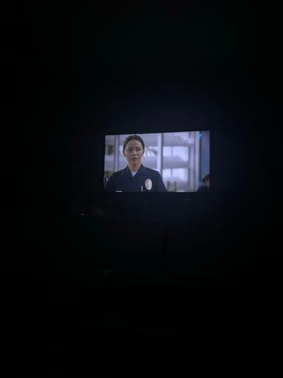 The image shows a television screen displaying a scene of a woman in a police uniform. The background appears to be an indoor setting, possibly a station or office. The room is dark except for the glow of the television.
There is no location information.