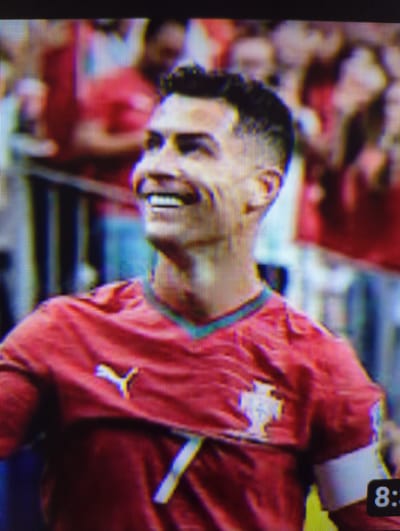 The image shows a close-up of Cristiano Ronaldo, a male soccer player, wearing a red jersey with a Puma logo on the left chest. The jersey also features a crest with a white shield and a stylized crown. He has short, dark, curly hair and is smiling broadly with his mouth open. The background is blurred and appears to be a stadium crowd, with many people in red clothing. The scene does not provide any specific information about the city of Erlangen, Germany.