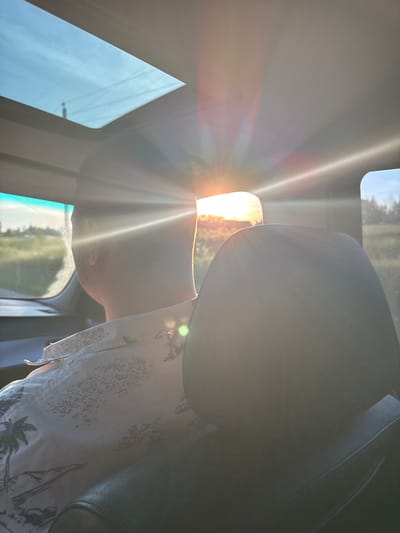 The image depicts the interior of a vehicle, presumably in motion, during a sunset or sunrise in Подчерково, Russia. The perspective is from behind the front passenger or driver's seat, looking forward. A male individual is seated, viewed from the rear, wearing a light-colored, short-sleeved collared shirt patterned with palm trees and tropical landscapes. His head and upper back are visible, with his hair appearing dark. A dark, possibly black, car seat headrest occupies the lower right foreground, partially obscuring the seated individual. Through the vehicle's windows, an intense, low sun is visible on the right, casting significant lens flares and light rays across the cabin, illuminating the person's right side and shoulder. Outside, a rural landscape with green fields and distant trees is visible. An open sunroof, revealing a clear blue sky with faint linear patterns, is present in the upper left of the vehicle's ceiling.