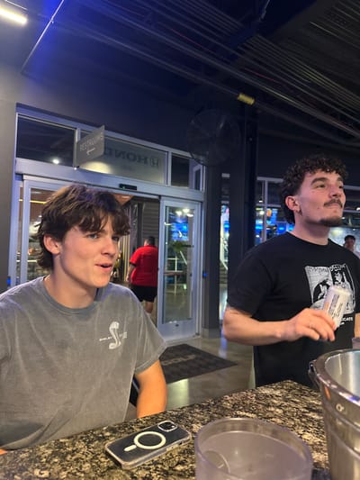 Two young men are seen standing near a high-top table with drinks and a phone. The person in the foreground is wearing a gray "Shelby Cobra" t-shirt, while the other person is wearing a black t-shirt. The background features the entrance to a building, possibly a Topgolf location, as a golf-branded floor mat is visible.