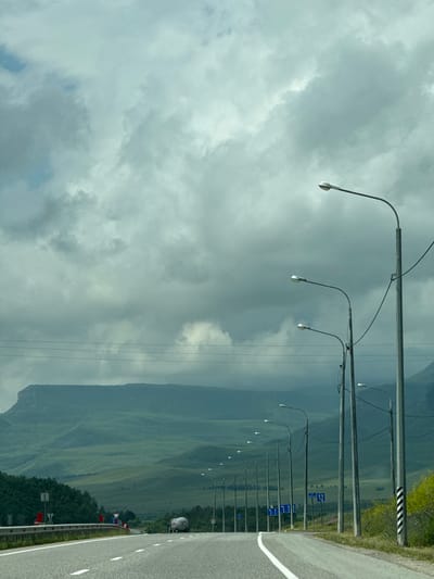 The image depicts a road scene on a cloudy day, likely on the outskirts of Kubina, Russia, given the presence of a road sign indicating "27" which could represent a route number or distance. The road, paved and divided by white lines, curves to the right and is flanked by a metal guardrail on the left. On the right side of the road, a series of streetlights are visible, extending into the distance. Beyond the road and streetlights, rolling green hills form the backdrop, with a prominent flat-topped mountain range in the far distance. Trees line the left side of the road, and a white tanker truck is visible further down the road. The sky is heavily overcast with gray clouds.