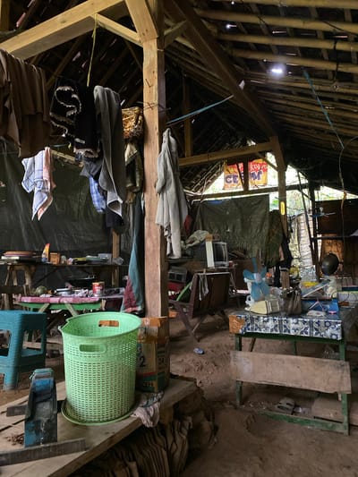 The image depicts the interior of a rustic, open-air structure, likely a workshop or storage area, located in Kresek, Indonesia. The building features a wooden beam and bamboo roof with gaps, allowing natural light to filter through. The floor is composed of dirt.

Various items are scattered throughout the space. Clothes are hanging on lines, some appearing to be laundry. Several tables, some constructed from rough wood and others with patterned tablecloths, are laden with objects including a fan, a television, cooking utensils, food items in packaging, and what appears to be a melon. A large green laundry basket is prominent in the foreground. There are also boxes, tarpaulins, and miscellaneous tools visible.

In the background, a person is partially visible standing near a ladder. The overall impression is one of a functional, somewhat cluttered, and lived-in environment.
