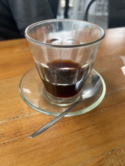 A close-up, eye-level shot shows a clear glass of dark liquid, likely coffee, sitting on a clear glass saucer. A silver spoon rests on the saucer to the right of the glass. The items are placed on a light brown wooden table with visible grain. In the background, slightly out of focus, a person is partially visible wearing dark clothing. The scene appears to be set in an indoor cafe or restaurant in Pulokiton, Indonesia.