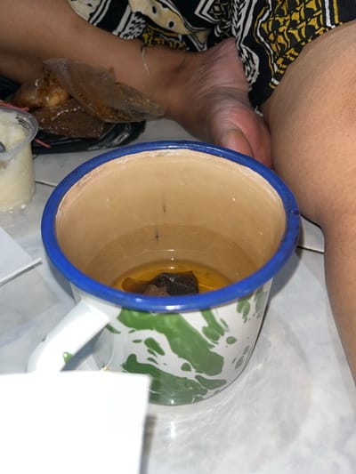 The image shows a close-up, high-angle view of a person relaxing indoors, surrounded by food and drink. A person's arm or leg is visible on the left side, covered partially by a black, yellow, and white patterned fabric. A foot is also discernible near the base of a prominent mug.

The central object is a white enamel mug featuring a blue rim and a distinctive green mottled pattern, resting on a light-colored tiled floor. Inside the mug, a small, dark object, possibly a tea bag or a piece of fruit, is partially submerged in a light brown liquid, suggesting a beverage like tea. The liquid level is low, indicating it has either been partially consumed or is in the early stages of preparation.

To the left of the mug, adjacent to the person, a clear plastic package contains a brown, somewhat translucent food item, which appears to be a snack or dessert. Below this, a portion of a small white plastic container with a spoon is visible, likely holding another food item.

The setting appears to be a casual indoor environment, typical of a home in Paweden, Indonesia, given the traditional enamel mug and tiled flooring. There is no visible text in the image. The time of day and the person's emotions are not discernible, but the scene conveys a sense of quiet relaxation.