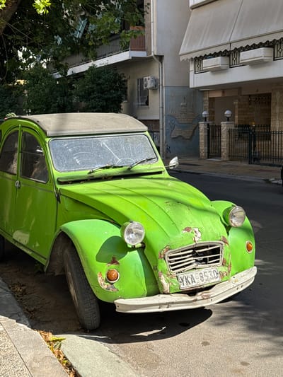 The image shows a bright green vintage Citroën 2CV parked on a street in Dafni, Greece. The car, which appears to be in a state of disrepair with peeling paint and rust, is the primary subject. The scene is an outdoor urban environment, with buildings and trees visible in the background. A sidewalk and a paved street are also part of the setting. The time of day appears to be daytime, with strong sunlight casting shadows. The weather seems to be clear. The car's license plate is visible and reads "KA 8530". The overall impression is one of a weathered, nostalgic artifact resting in a typical Greek streetscape.