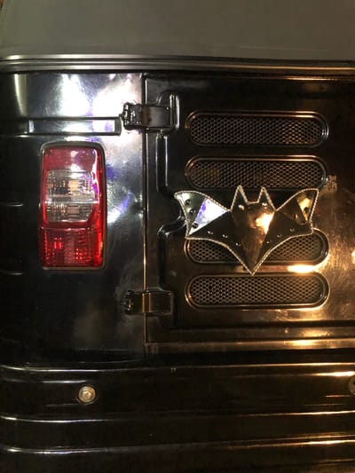 The image shows a close-up view of the rear of a black vehicle, possibly a jeep or SUV, with a Batman logo affixed to the tailgate. The vehicle's taillight, a red and clear rectangular unit, is visible on the left side of the image. The Batman logo, a metallic bat shape with pointed ears and studded detailing along its border, is centered on the tailgate, positioned between two horizontal, oval-shaped grilles. The vehicle's surface is black and appears to be highly polished, reflecting light sources that create bright highlights and shadows. A visible bolt or screw is located on the lower section of the tailgate. The environment appears to be indoors or in a dimly lit area, with artificial lighting contributing to the reflections on the vehicle's surface. There is no indication of the location being Karandugoda, Sri Lanka, as the image focuses solely on the vehicle's rear.