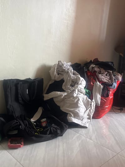 In a room in Serdang Kulon, Indonesia, a pile of clothes is visible on the floor. There is a large red plastic bag filled with clothing, and a black tote bag. Several pieces of clothing are strewn on the floor, including a white t-shirt and black pants. A pack of cigarettes and an ashtray are also on the floor. The walls are painted a light color, and the floor is tiled.