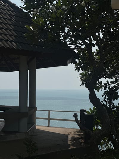 The image shows an outdoor scene, likely a balcony or veranda, overlooking a body of water. The view is partially framed by a tree with dense foliage at the top. The structure of the balcony consists of a dark tiled roof section in the lower left, white support beams, and a grey concrete railing. The body of water is slightly hazy. This image was taken in Ao Hin Khok, Thailand.