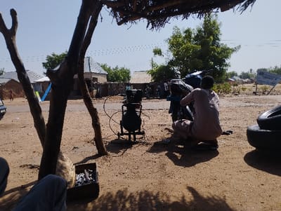 The image shows a man sitting on the ground in front of a motorcycle in Tongo, Nigeria, at the coordinates (latitude: 10.664, longitude: 11.364). The man is wearing a long-sleeved shirt and pants, and he is working on the motorcycle's tire. There is a tire iron next to him, as well as an air compressor. The man is sitting in the shade of a tree, which is growing next to a small, dirt road. There are other buildings and trees in the background. The man's back is to the camera. The sky is clear and the sun is shining. The motorcycle is parked in front of a building with a blue roof. The building's façade is partially obscured by the tree's branches. A wooden box sits on the ground next to a tree trunk. The scene is a typical example of a street mechanic working on a motorcycle in Tongo, Nigeria.