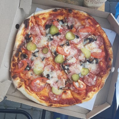 A close-up, top-down shot captures a half-eaten pizza in a cardboard pizza box. The pizza, with a golden-brown crust, is topped with melted cheese, sliced salami, mushrooms, tomatoes, and pickles. The pizza box is open, revealing the contents and a white paper liner. The background shows a wooden surface, and a portion of a paper cup with faded text is visible. The overall scene is set outdoors, with hints of a paved surface and greenery suggesting a possible urban or suburban environment in Dubrivka, Ukraine.