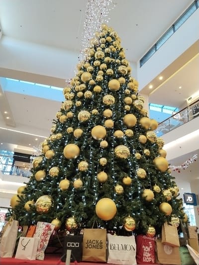 The scene depicts a large, artificial Christmas tree centrally positioned within the multi-level atrium of the Dos Mares Shopping Centre in San Javier, Spain. The tree is densely adorned with numerous spherical ornaments of varying sizes, exhibiting both matte and reflective gold finishes, and is illuminated by strings of small white LED lights. At the base of the tree, an arrangement of shopping bags from various retailers, including "JACK & JONES," "BELMON," a black bag with three white squares, and a red bag with "HOP HOP HOP" text, serves as decorative elements, simulating wrapped gifts. The background reveals the shopping center's modern interior architecture, featuring light-colored walls, high ceilings with recessed lighting, and upper-level walkways equipped with glass and metal railings. Store signage for "JD" is visible on an upper floor. Delicate white and red string lights are suspended from the ceiling above the tree. No individuals are present within the captured frame.