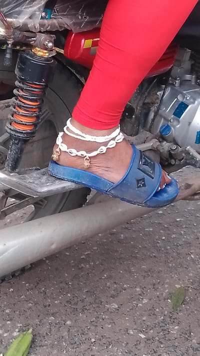 A close-up shot focuses on a person's left foot wearing a blue slide sandal with a "LV" logo. The foot is adorned with a white anklet made of shells and small golden charms. The person is wearing red leggings, and their foot is resting on a metal part of what appears to be a motorcycle. The background shows parts of a motorcycle, including a shock absorber and engine components, and a grey, gravelly surface that is consistent with a street in Agbarho, Nigeria.