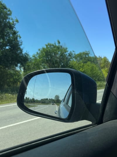 The image is a close-up, POV shot from inside a vehicle traveling on a road with green trees on either side. A side-view mirror reflects the road behind, showing a clear blue sky and a single car in the distance. The scene appears to be in a rural or suburban area, not specifically within the city of Ленинское, Russia. There are no people visible, and the action is limited to the car's movement and the reflection in the mirror. The environment is characterized by a paved road, vegetation, and a bright, sunny day.