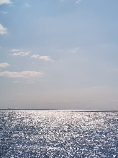 The image depicts a wide expanse of water, likely a sea or large lake, shimmering under a bright sky. The water's surface is highly textured with countless small waves catching the sunlight, creating a dazzling, almost glittery effect. A thin, hazy horizon line separates the water from the sky, with faint landmass features or distant structures barely discernible. The sky is a pale blue, populated by scattered, wispy clouds. The overall impression is one of open space and natural light. There are no people, animals, or man-made objects clearly visible in the scene, giving it a tranquil and untouched feel. The location is not specified in the image.