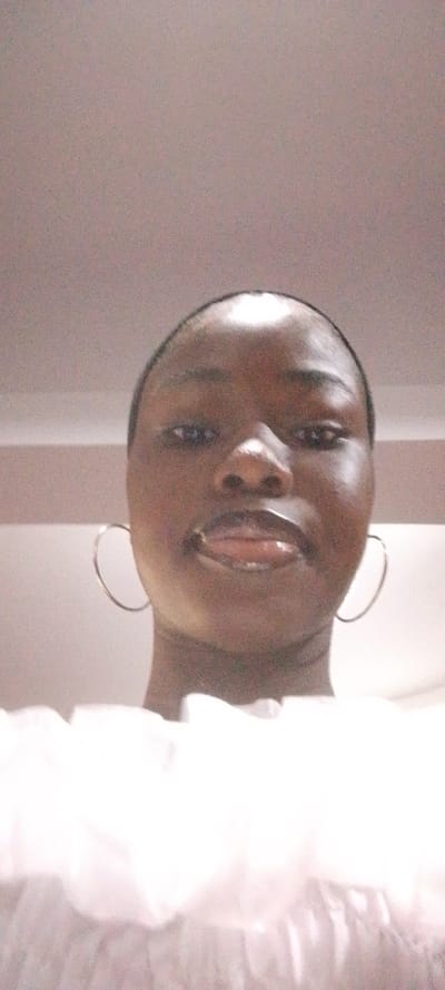 A close-up, low-angle portrait features a dark-skinned individual, likely female, positioned centrally against a plain, light-colored interior background. The subject's face dominates the frame, showcasing a broad forehead, dark eyes looking directly forward, a slightly upturned nose, and full, glossy lips parted slightly. Dark hair appears pulled back or short. The individual wears large, silver-colored hoop earrings. A white garment with distinct ruffles or gathered fabric is visible across the bottom third of the image, covering the chest area. The background consists of a uniform light-toned ceiling and upper wall, indicating an indoor setting with ambient artificial lighting. The specific location within the city of Ohokobe Ndume, Nigeria, is not discernible from the visual evidence in the image.
