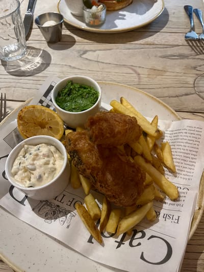 The image displays a close-up, eye-level view of a meal served at a restaurant or pub in the Borough of Runnymede, United Kingdom. The central subject is a serving of fish and chips presented on a speckled ceramic plate lined with newspaper-style paper.
The meal consists of two pieces of battered, deep-fried fish, accompanied by a generous portion of golden-brown french fries. Two small white ramekins are present: one contains a vibrant green substance, identified as mushy peas, and the other holds a creamy white tartar sauce with visible relish pieces. A charred half-lemon is placed next to the ramekins. The newspaper-style paper under the food features text, including "Fish and chips," "UK fish and chip shops," and a crest-like logo with "MON.REP. EST. 1852 LUXE" and illustrations of fish.
The immediate dining environment shows a light-colored wooden plank table. To the left, a clear, textured drinking glass filled with water, a silver knife, and a small cylindrical metal cup are visible. In the background, partially visible elements suggest another place setting or additional items, including a white plate with what appears to be a burger, a small glass dish with condiments, two silver forks, and the stem of a wine glass. A faint reddish stain is present on the wooden table surface. No individuals are visible in the frame.