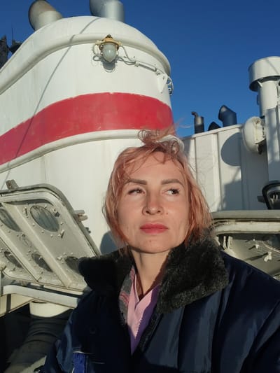 A medium shot photo shows a woman with short, pinkish-blonde hair standing in front of a white, cylindrical structure with a red stripe. The woman is looking up and to her right, with her mouth slightly open. She is wearing a dark blue jacket with a fur collar and a pink shirt. The background shows a bright blue sky and parts of a large white object, possibly a ship or industrial equipment, with pipes and a light fixture visible. The image appears to be taken outdoors during the day.