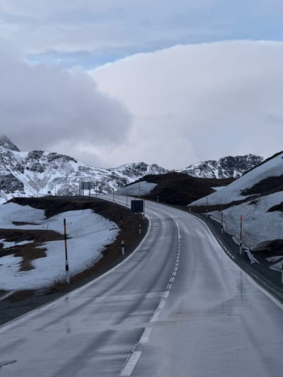 The image, depicting a high-altitude mountain pass, presents a winding, wet paved road under an overcast sky. When viewed in its correct landscape orientation (rotated 90 degrees counter-clockwise from the provided image), the road stretches into the distance, characterized by clear white dashed and solid lane markings.

Flanking the road are steep, rugged mountains with dark, exposed rock and significant patches of residual snow, indicative of a high alpine environment. Numerous road markers, consisting of black and white posts with red or black tips, line the road's edges, likely for visibility in snowy conditions. A few small, dark rectangular signs are visible beside the road, but no discernible text is legible on them.

The sky is predominantly covered by thick, diffused gray clouds, with a small section of lighter blue sky visible near the horizon. The wet road surface reflects the soft, diffused light, contributing to the stark yet majestic atmosphere. The scene is devoid of people or vehicles, conveying a sense of solitude and the raw grandeur of the mountain landscape, consistent with a pass in a region like Poschiavo, Switzerland. The time of day appears to be daytime, under cloudy conditions.