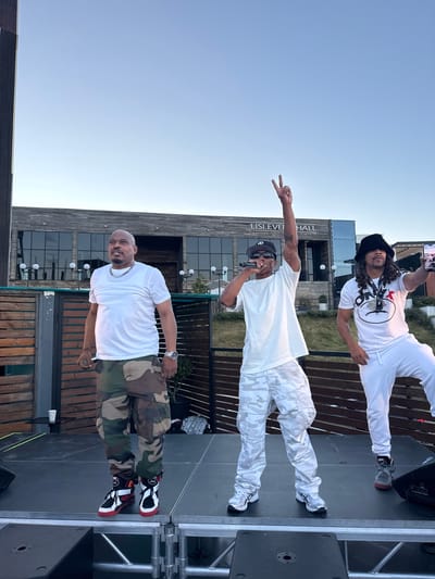 Three men are standing on a stage in front of a building with "LISI EVENT HALL" signage. The man on the left is wearing a white t-shirt, camouflage cargo pants, and red and white sneakers. He is looking to his right. The man in the center is wearing a white t-shirt, white camouflage cargo pants, and white sneakers. He is holding a microphone and has his left arm raised with two fingers up. He is looking to his right. The man on the right is wearing a white t-shirt with a logo, white pants, and gray sneakers. He is holding a phone in his right hand and looking at the phone. The background shows a modern building with large windows and an outdoor seating area with wooden fences and tables. The sky is clear and blue. The image appears to be taken outdoors during daylight. There is no information to indicate the location is in Mukhatskaro, Georgia.