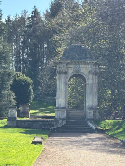 The image depicts a stone pavilion or folly with a domed roof, located in a garden setting, likely within the Malvern Hills area of the United Kingdom. The structure is built of light-colored stone, with an arched opening and decorative details. It is positioned at the top of a set of stone steps leading down into the garden. The scene includes trees in the background and manicured lawns. The photograph appears to be taken on a sunny day. There are no people visible in the image.