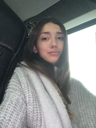 The image features a young woman inside what appears to be a car interior, captured with the frame rotated such that her head is on the left side. She has long, dark brown hair, light brown eyes, and full lips, looking directly at the camera with a calm and neutral expression. She is wearing a light grey, thick knit sweater or a similar warm garment.
The setting indicates she is likely a passenger in a vehicle. To the left of her head, parts of a black car seat are visible, along with some circular elements that could be car controls or air vents. Above her, a large, bright white area dominates the upper right portion of the frame, consistent with a window or sunroof letting in bright daylight. The strong, diffused light suggests an overcast sky or bright ambient light conditions.
The woman is depicted in a reclining or resting posture. No specific activity other than posing for the photograph is evident. There is no visible text within the image. The general location context for this scene is Stara Pazova Municipality, Serbia. The overall impression is relaxed and casual.