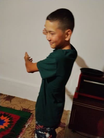 In Almaluu, Kyrgyzstan, a young boy with short dark hair and wearing a dark green t-shirt and patterned pants stands with his back to a plain white wall. His right arm is bent at the elbow and extended outwards, with his hand open and facing away from his body. He is smiling and looking to his right. In the background, a dark pot sits on top of a dark red piece of furniture with drawers. The floor is covered by a rug with an intricate geometric pattern.