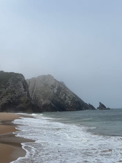 The image depicts Praia da Adraga, a beach in the municipality of Colares, Portugal. The scene is dominated by a rugged coastline with steep, rocky cliffs that are partially covered in vegetation. The sea is a moderate blue-gray with white, foamy waves breaking on the shore. The beach itself is composed of light brown sand. The sky is overcast, creating a diffused light and a somewhat muted color palette. In the distance, there are several sea stacks rising from the water. A lone figure, appearing as a dark silhouette, is visible walking on the beach in the far left of the image.