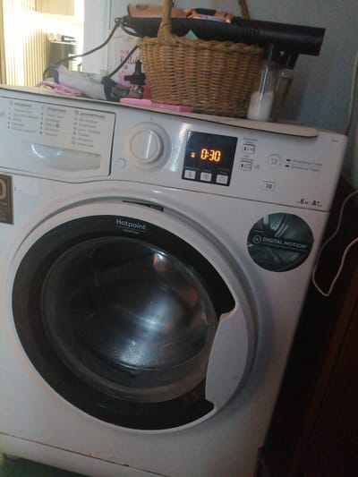 A white Hotpoint Ariston washing machine is shown in a room. The machine has a digital display showing "0:30". There is a basket on top of the machine, and various cleaning supplies. The room appears to be a laundry room or bathroom. The image is taken from a slightly elevated angle, looking down at the washing machine. The lighting is dim, with some light coming from a window to the left of the frame. The overall impression is of a functional, but somewhat cluttered, domestic space.
