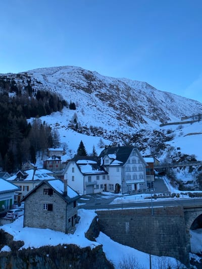 The image displays a panoramic view of the village of Hospental, Switzerland, during winter. Snow covers the ground, rooftops, and the surrounding mountainous terrain under a clear, pale blue sky.
In the background, a large snow-covered mountain dominates, featuring sparse dark coniferous trees on its lower slopes and visible rock formations. Avalanche protection netting or terracing is visible on the upper left slope of the mountain.
The village occupies the mid-ground, nestled in a valley. Prominent structures include a multi-story, light-colored building with numerous windows and decorative white trim, identified as "GASHAUS ROGGENRUDD," possessing an outdoor patio with white tables and chairs. Adjacent to it are smaller, white-plastered buildings with dark roofs. In the foreground, positioned on a snow-covered bank, is a two-story house constructed of light-colored stone with a dark roof and small windows, some with closed shutters. Other residential buildings with varied facades and dark or red roofs are scattered across the snow-laden landscape, extending up the gentle incline.
A dark asphalt road winds through the village, partially cleared of snow. A stone-arch bridge supports a road or railway track over a ravine on the right side of the image, with snow accumulating on its banks and surrounding areas. Another, lighter-colored bridge is visible further into the valley. A single white car is parked near a building on the left. No individuals or active interactions are visible. The scene is illuminated by soft, cool natural light, casting minimal shadows.