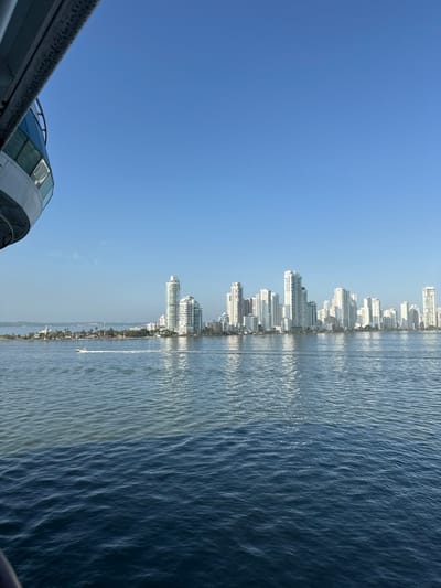 The image displays a panoramic view of the Bocagrande skyline in Cartagena, Colombia, as seen from a vessel navigating the Bahia de Cartagena. The upper-left quadrant reveals a partial view of a ship's structure, including a white railing and blue-tinted windows. The foreground and midground consist of water, which presents two distinct color zones: a darker blue in the immediate vicinity of the observer, transitioning to a lighter, somewhat turbid blue-green closer to the distant landmass. Ripples are visible across the water surface. A white speedboat is observed in motion from right to left across the lighter water, leaving a visible wake. To the left of the main urban area, a low-lying, vegetated landmass or island is present, featuring a small, light-colored structure resembling a lighthouse on its western tip. The background is dominated by a dense cluster of numerous modern high-rise buildings, predominantly white or light-colored, forming the characteristic skyline of the Bocagrande peninsula. The sky above is clear and uniformly blue, devoid of clouds. No specific individuals are discernible.