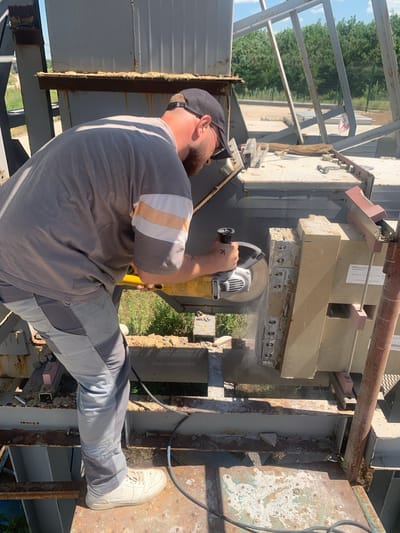 A man wearing a grey t-shirt with striped sleeves, a black baseball cap, and grey work pants is using an angle grinder to cut a beige concrete block. He is standing on a metal structure outdoors, likely a construction site, given the presence of scaffolding and the unfinished nature of the surroundings. The sun is shining brightly, casting distinct shadows, suggesting it is daytime. The man appears focused on his task, with no discernible emotion. Dust is being generated from the grinding process. In the background, trees and a fence are visible, hinting at a rural or suburban setting. The concrete block he is cutting is supported by metal brackets and bolted to a larger structure. There is some visible rust on the metal components of the structure. A label is partially visible on the concrete block. The image is oriented vertically. The location is identified as Pankratove, Ukraine.