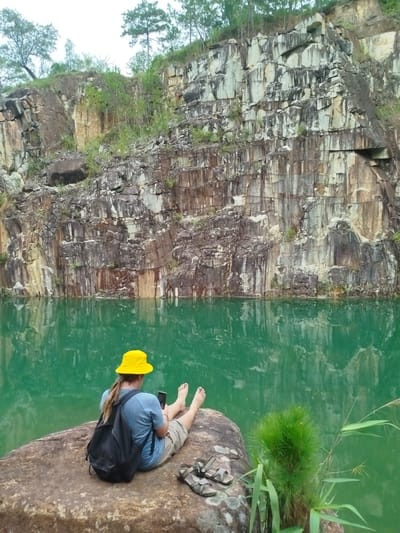 A person is sitting on a large rock by a body of water. The person is wearing a yellow bucket hat, a blue t-shirt, and tan shorts. A black backpack is on the person's back. The person is holding a mobile phone and looking at it. The person's feet are visible and bare. A pair of sandals are on the rock next to the person. The water is green and reflects the surrounding cliffs. The cliffs are made of layered rock with some vegetation growing on them. The sky is overcast. The scene appears to be a natural setting, possibly a quarry, in Lán Tranh, Vietnam.